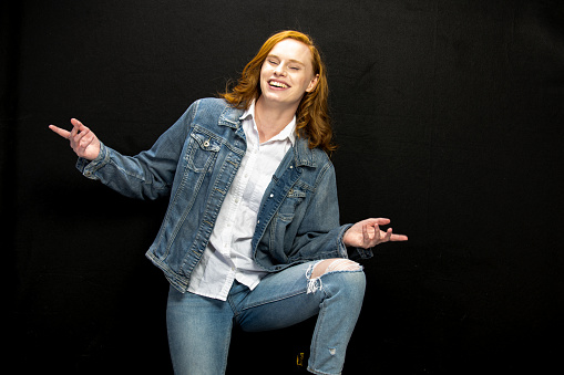 Young Los Angeles citizen with long red hair and dressed in a white shirt and denim outfit poses in studio before a black backgound