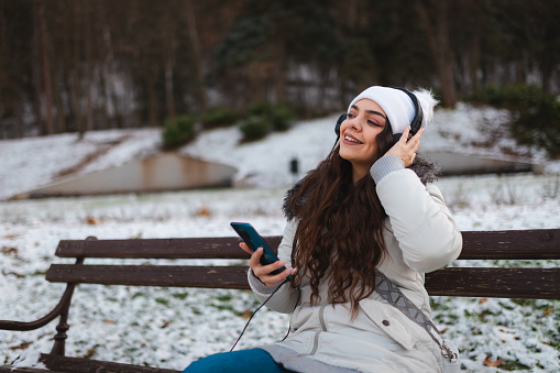 Young woman listening to music outdoors while wearing warm clothing. It is wintertime.