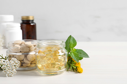 Jars with different pills, flowers and herbs on white wooden table, space for text. Dietary supplements