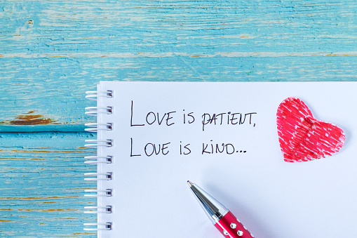 Love is patient and kind, handwritten verse in notebook with pen and red paper heart on wood. Christian biblical concepts of care, comfort, compassion, grace, and service.