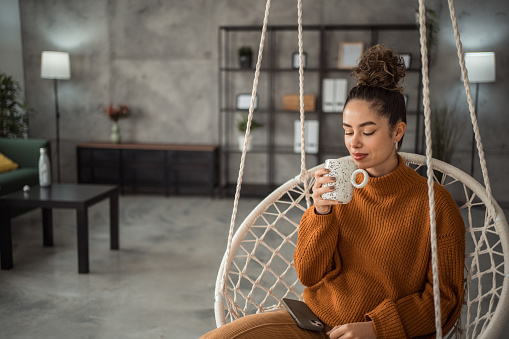 A beautiful young woman is relaxing and drinking coffee while swinging in a swing in the living room