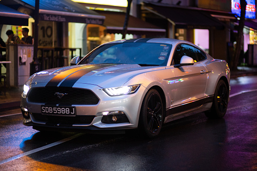 A Ford Mustang drives through the neon lit roads on a rainy evening during monsoon season, Singapore