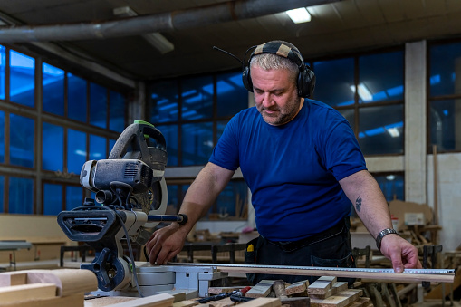 A middle-aged carpenter works on a table circular saw in the carpentry workshop.