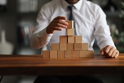 Businessman hand pulling out or placing wood block on the tower and semi-pyramid in modern office. Plan and strategy in business.