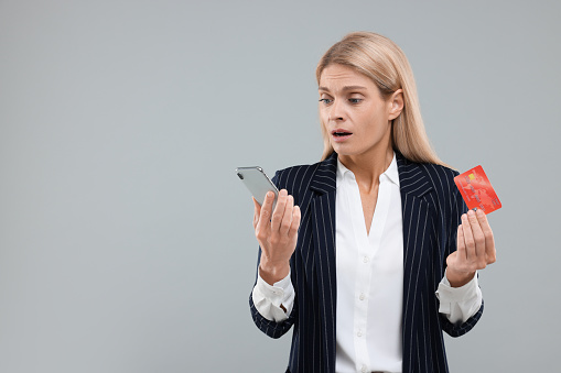 Stressed woman with credit card and smartphone on grey background, space for text. Be careful - fraud