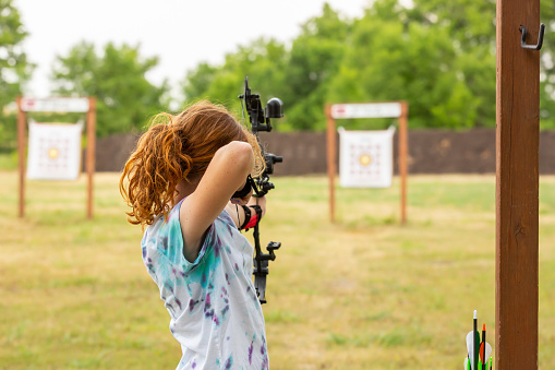 Rear view of a girl practicing archery at an outdoor range on an overcast summer day. The girl has her bow drawn and is aiming at the target in the distance.