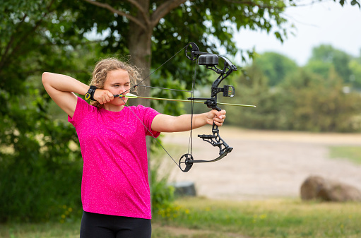 Close-up of a teenage girl practicing archery at an outdoor range on a summer day.