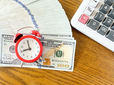 Alarm clock on top of 100 dollar bills with calculator on a wooden desk