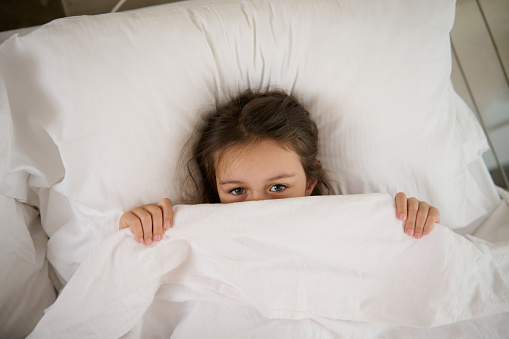 Little girl lying on the bed, hiding half of her face under a white blanket, looking at camera. Surprised child peeking from duvet wake from bad dream sleep feel fear afraid of nightmare, top view