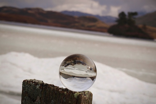 View over a frozen mountain lake with snow and mountains in the background with focus on a glass lens ball on a wooden post in the foreground