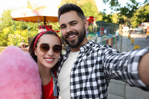 Happy young man and his girlfriend with cotton candy taking selfie at funfair