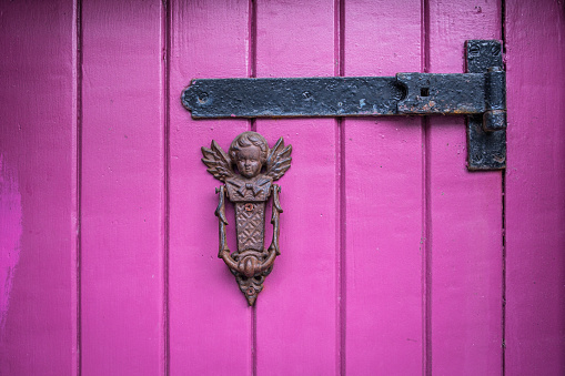 An old bronze door knocker in the shape of an angel, on a pink wood paneled door with a black metal hinge.