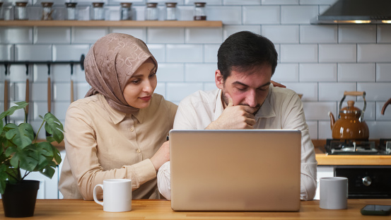 Married couple sitting in the kitchen using laptop saddened by the news, the loss, the wife consoles the husband. Disappointed bad news, loss negative result