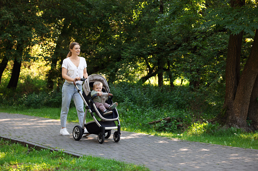 Happy nanny with cute little boy in stroller walking in park, space for text
