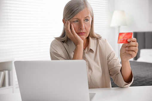 Upset woman with credit card near laptop at table indoors. Be careful - fraud