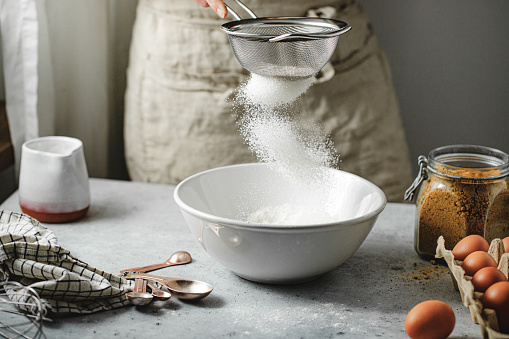 Cropped shot of a woman making pancake in the kitchen. Hand of a female sifting the flour with a steel sieve in a ceramic bowl with other ingredients on kitchen table.