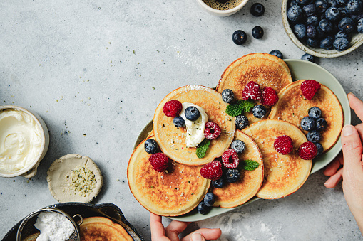 Close-up of a woman hands serving freshly prepared pancakes on table. Female placing stack of homemade pancakes with berries on a tray.