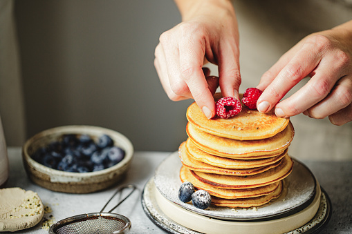 Close-up of a woman serving healthy breakfast. Female hands garnishing pancake stack with raspberries and blueberries on kitchen table.