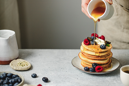 Close-up of woman pouring maple syrup over homemade pancakes. Female serving homemade pancakes on breakfast table.