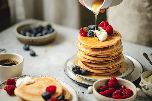 Close-up of woman pouring maple syrup over stack of pancakes over dining table. Woman serving homemade pancakes on breakfast table.