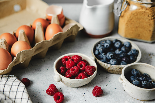 Close-up of food ingredients on kitchen counter for preparing healthy breakfast. Brown eggs in a carton with raspberry and blueberries in bowls on kitchen table.