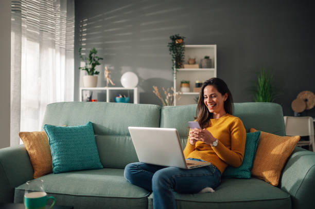 istock Woman using laptop and typing on a phone while sitting on couch at home 1938209173