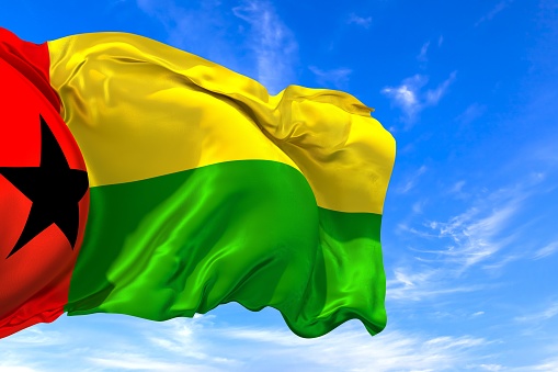 The national flag of Guinea-Bissau with fabric texture waving in the wind on a blue sky. 3D Illustration