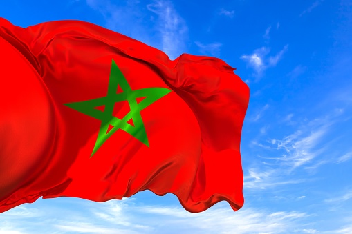 The national flag of Morocco with fabric texture waving in the wind on a blue sky. 3D Illustration