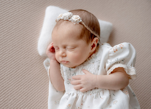 Baby Girl In Dress Sleeps On Beige Fabric Background During Newborn Photo Session In Studio