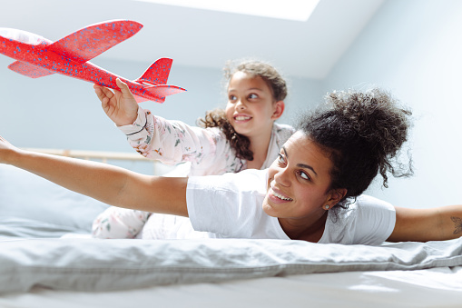 Mother and daughter paying in bedroom in the morning. Little girl is holding airplane and having fun on relaxed weekend morning