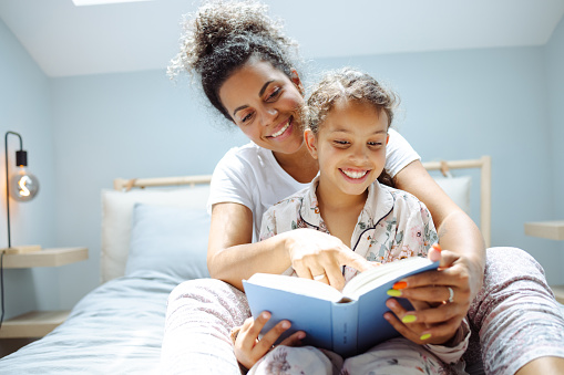 Latin mother smiling and reading book in bedroom to her cute daughter. They are enjoying in bedroom during weekend