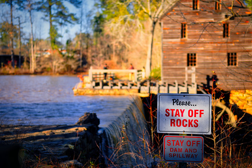 A fall landscape at sunset of warning signs at the dam's slippery rocks at Yates Mill Park downstream in Raleigh, North Carolina.