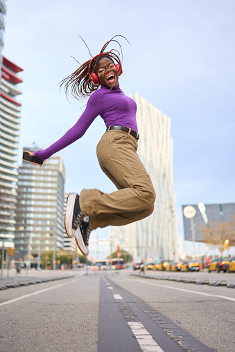 An ecstatic young woman with vitiligo and braids leaps high, listening to music in the heart of the city