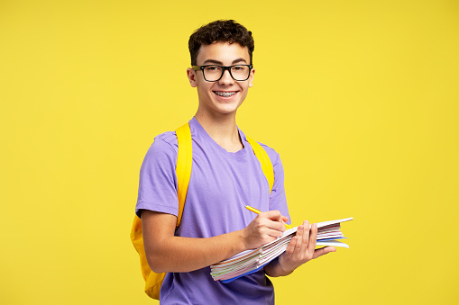 Smart smiling school boy wearing eyeglasses holding books taking notes isolated on yellow background. College student with backpack studying, learning language looking at camera. Education concept