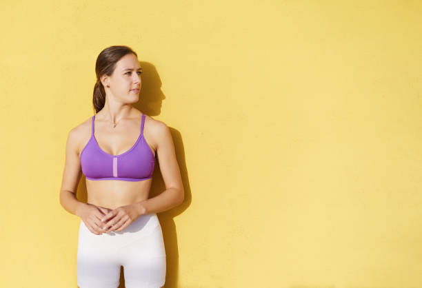 Young athletic female in purple top and white shorts standing against yellow background with shadow and looking away