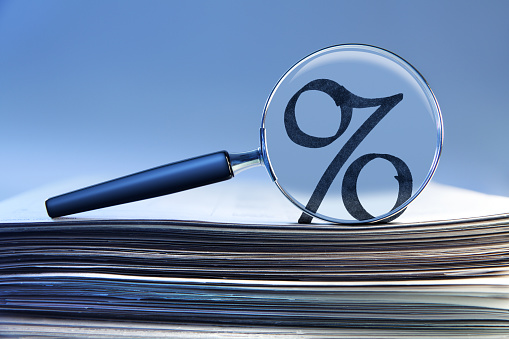 A percentage sign seen through a magnifying glass that rests on top of a stack of financial documents.