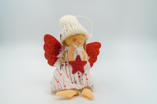 Handmade knitted Christmas angel doll, red glitter wings, holding red star, adorned with white hat, blond yarn hair, heart-printed dress, golden feet, smiling wooden face. White background