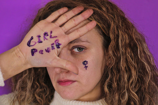 Pretty white young girl with curly hair with hand covering half her face, showing her blue eyes, and with representations of feminism drawn on her hands, purple background. Concept of revolution