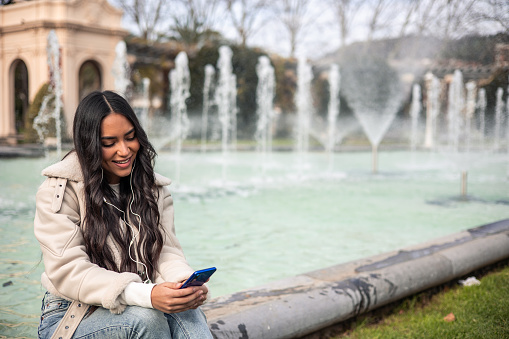 young latin girl smiling with headphones and cell phone sitting in a city fountain