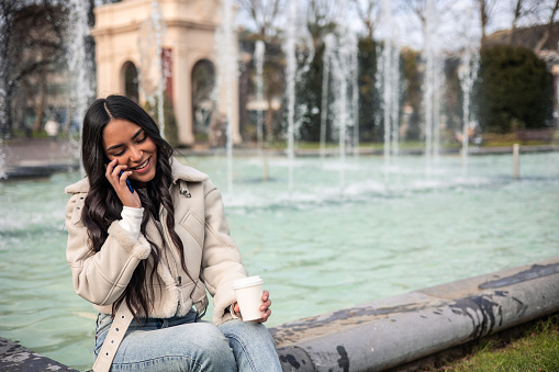 young latin girl making a call with the cell phone in her right hand and a coffee in the other sitting at the fountain in the city