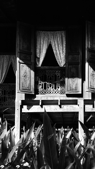 A black and white photo of front portion of a typical traditional Malay house . The main part is the large open windows with decorative art on its doors and fence.