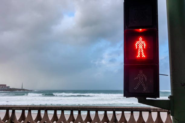 This thought-provoking photograph juxtaposes the rigid control of a traffic signal with the untamed force of the sea. The red silhouette of a pedestrian is illuminated in a clear command to stop, set against a backdrop of wild, churning waves and a stormy sky. The balustrade runs parallel to the horizon, while the traffic light stands as a solitary figure of human imposition. The contrast is striking the controlled, static world of man-made objects against the ever-moving, dynamic nature of the ocean.