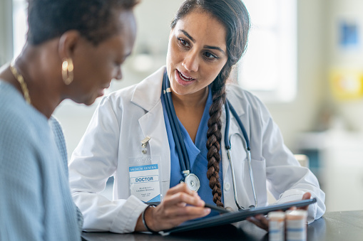 A senior woman of African decent meets with her doctor as they review her medications together.  The doctor is dressed professionally in scrubs and a lab coat and is holding out a tablet between the two as they review her recent test results together.