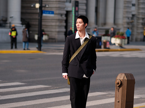 Portrait of handsome Chinese young man with black short hair wearing black blazer standing on street with modern city building background in sunny winter day, male fashion, cool Asian young man.