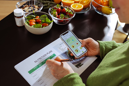 A female nutritionist checks the nutrition facts and calorie intake of the meals on her smart phone. She is writing a nutrition plan for a client