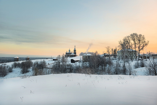 St. Elijah's Church and Trinity hill in Cherdyn. Ancient architecture is surrounded by tall white drifts and orange sunset skies. Historical center of the ancient city of Cherdyn, Russia of winter.