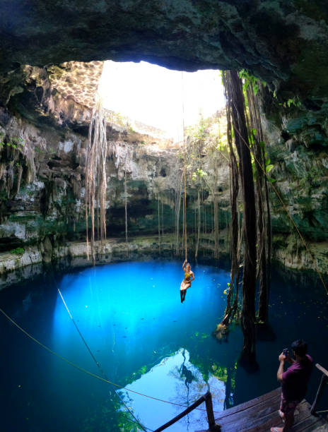 Valladolid, Mexico - April 15, 2021: Woman swings from a ceiba tree root hanging into the open cenote Oxman on a spring day