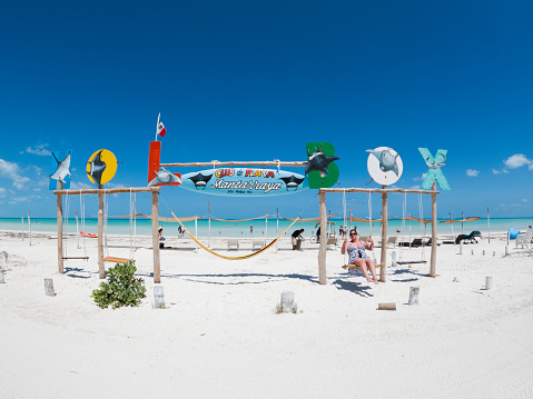 Holbox Isle, Yucatan, Mexico - April 8, 2021: Female tourist swings on the beach under a welcome sign for Manta Raya