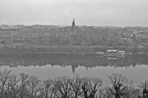 View of Georgetown University from across the Potomac river.   Cloudy, cold sky in the winter.   Boat house.  Black and white photograph.   Reflection of buildings on water.
