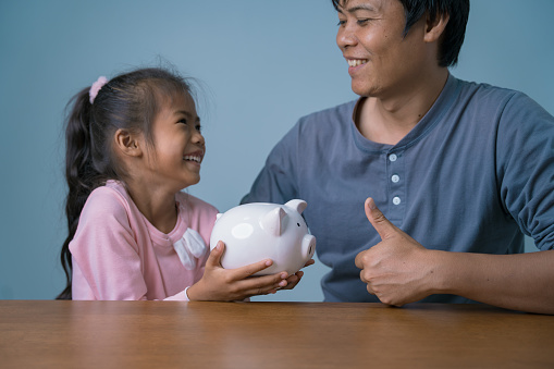 Asian man and daughter with a piggy bank, happy smiling faces. Idea of father and child helping each other put coins in a piggy bank to save money for scholarships or other expenses.